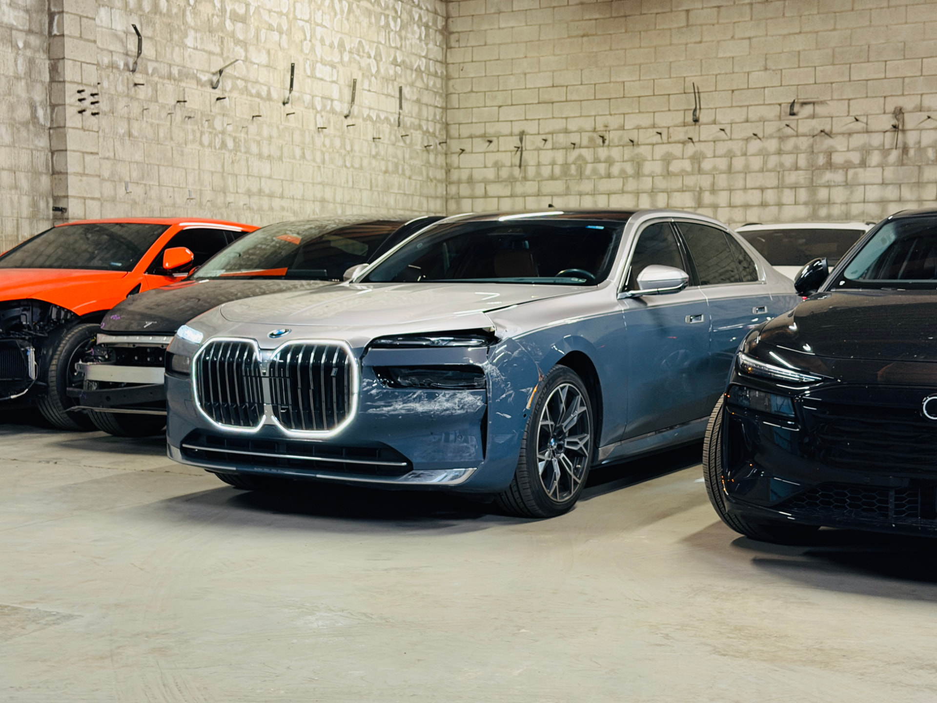 A damaged blue BMW 7 Series with a torn front wing, waiting for bodywork among other cars inside the shop