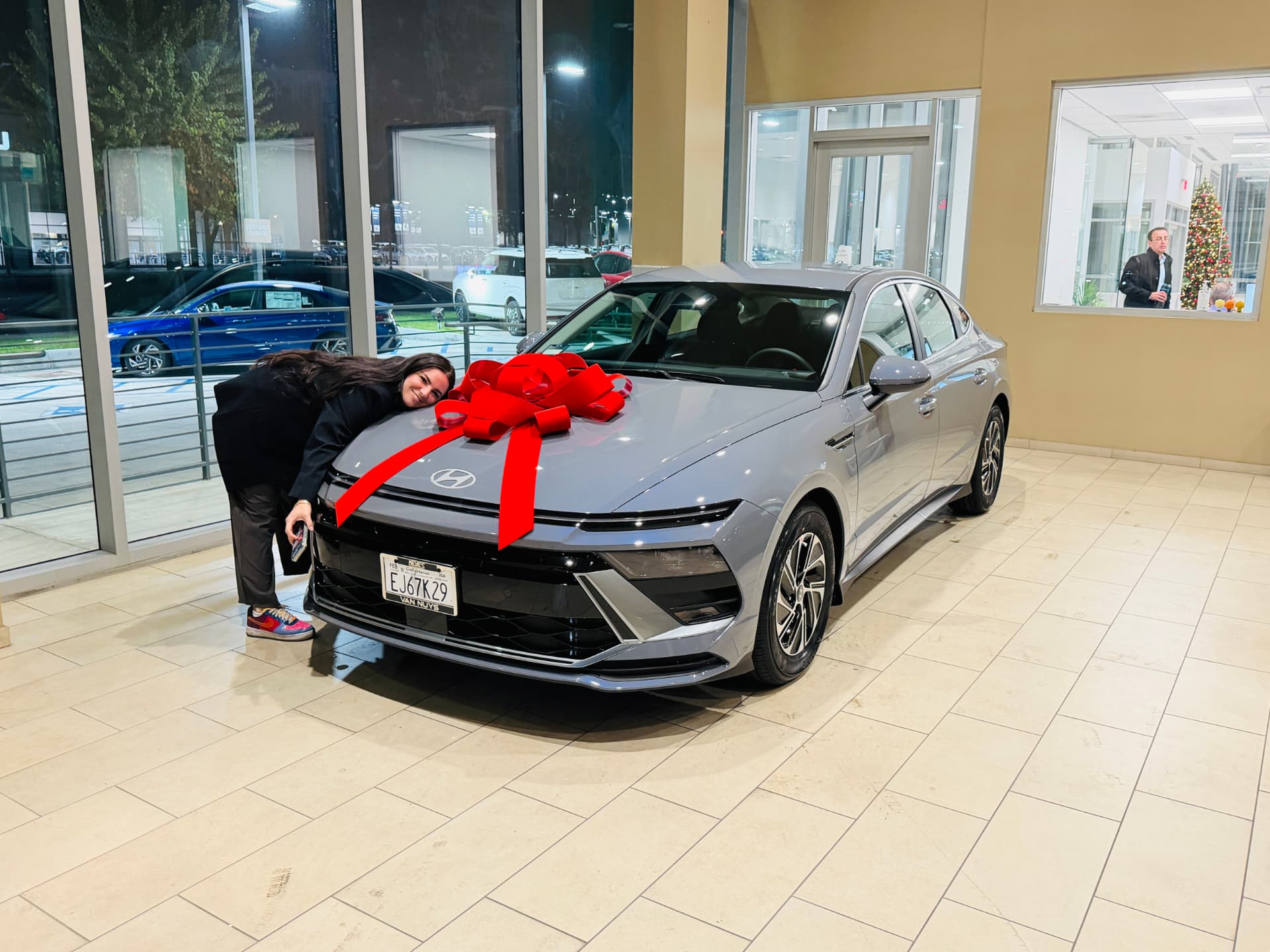 A customer leaning on the hood of a brand-new silver Hyundai Sonata with a red gift bow in a dealership showroom